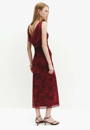 Mujer que lleva un vestido midi sin mangas, de flores en rojo intenso, con espalda drapeada y tirantes finos, combinado con tacones negros de tiras y pulseras doradas.