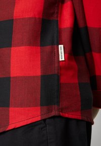 Camisa de franela a cuadros rojos y negros con una etiqueta en el lateral que dice "REDEFINED REBEL". Presenta un ajuste relajado con un dobladillo visible.