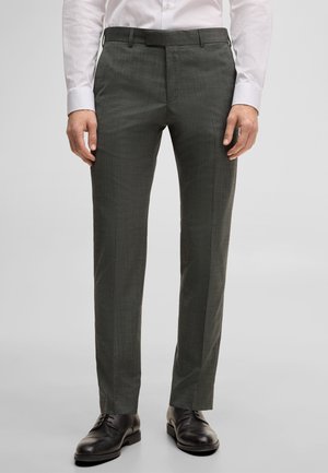 Man draagt op maat gemaakte donkergrijze nette broek met een wit overhemd en zwarte leren nette schoenen, staand met handen ontspannen langs het lichaam.