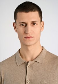 Beige stickad polo skjorta med korta ärmar, ribbad krage och två knappar fram. Har en diskret logotypdetalj på vänster bröst.