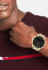Montre dorée avec un cadran noir et des accents argentés, dotée de trois sous-cadrans, portée sur un poignet marron, à côté d'un pull bordeaux.