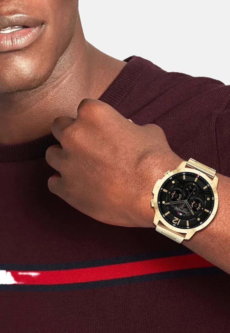 Montre dorée avec un cadran noir et des accents argentés, dotée de trois sous-cadrans, portée sur un poignet marron, à côté d'un pull bordeaux.