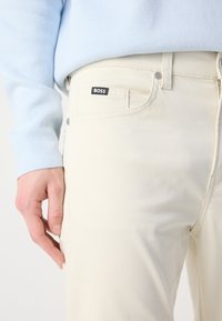 Lichtgekleurde jeans met een slim fit, voorzien van een zwart "BOSS" label op de zak, vergezeld door een zachtblauwe trui.