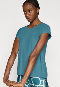 Μπλε-πράσινη αθλητική μπλούζα κοντομάνικη από απαλό ύφασμα· στρογγυλή λαιμόκοψη και άνετη εφαρμογή. Διαθέτει μικρό λογότυπο κοντά στον ώμο.