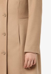 Cappotto in lana beige con cinque bottoni, una tasca laterale nascosta e una texture liscia con una vestibilità su misura, maniche accorciate.