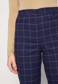 Beige långärmad topp instoppad i navyblå rutiga byxor med knapp och bältesöglor, hand vilande vid sidan.