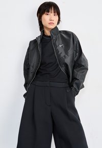 Casaco refletor preto com gola alta, bolsos laterais e fecho éclair. Usado sobre um top preto texturizado e calças largas pretas.