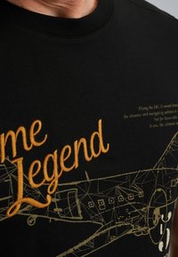Czarna bawełniana koszulka z okrągłym dekoltem, z złotym haftowanym napisem "Legend" oraz stylizowaną grafiką samolotu w złotej linii.