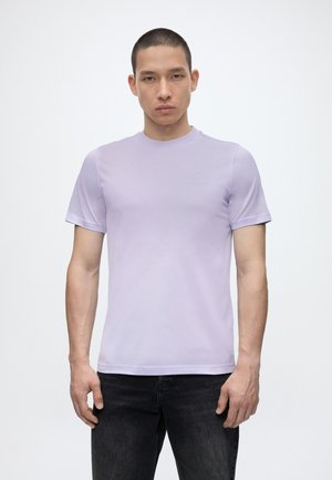 Młody mężczyzna o krótkich włosach, ubrany w gładki jasnofioletowy t-shirt z krótkim rękawem i czarne jeansy, stojący na białym tle.