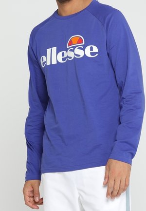 Uomo che indossa una camicia a maniche lunghe blu con un grande logo "ellesse" bianco e arancione, abbinata a pantaloni bianchi.