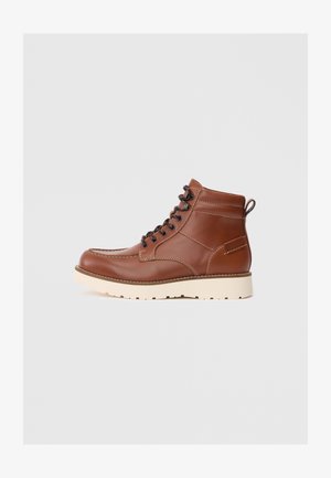 Stivaletti in pelle marrone con una texture liscia, suola in gomma bianca e lacci neri. Presentano una linguetta sul retro e cuciture a contrasto.