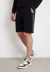 Pantalones cortos de sudor negro con bolsillos laterales, que presentan un logotipo blanco a los lados. Combinados con zapatillas gris y blancas, de pie sobre una superficie gris clara.