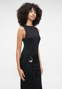 Μαύρο αμάνικο φόρεμα με εφαρμοστή σιλουέτα και ραμμένα υφάσματα, διαθέτοντας έναν κυκλικό ασημένιο διακοσμητικό στοιχείο στη μέση. Λείος υφασμάτινος απόχρωση.