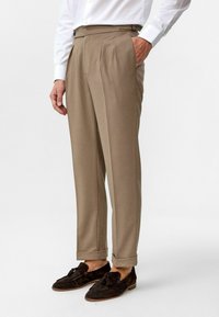 Beige geplooide broeken van een gestructureerde stof, met een hoge taille en opgerolde manchetten, gecombineerd met donkerbruin suède loafers.