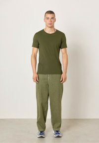 Olivově zelené bavlněné tričko s krátkými rukávy, kombinované se světlými olivovými volnými kalhotami a modrými sportovními botami na neutrálním pozadí.