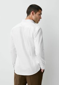 Weißes Langarmhemd mit spitzem Kragen, das eine glatte Textur und ein gerades Rücken-Design aufweist, kombiniert mit braunen Hosen.