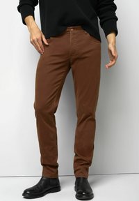 Man in bruin slim-fit broek en zwarte leren enkelboots, staand tegen een effen lichte achtergrond.