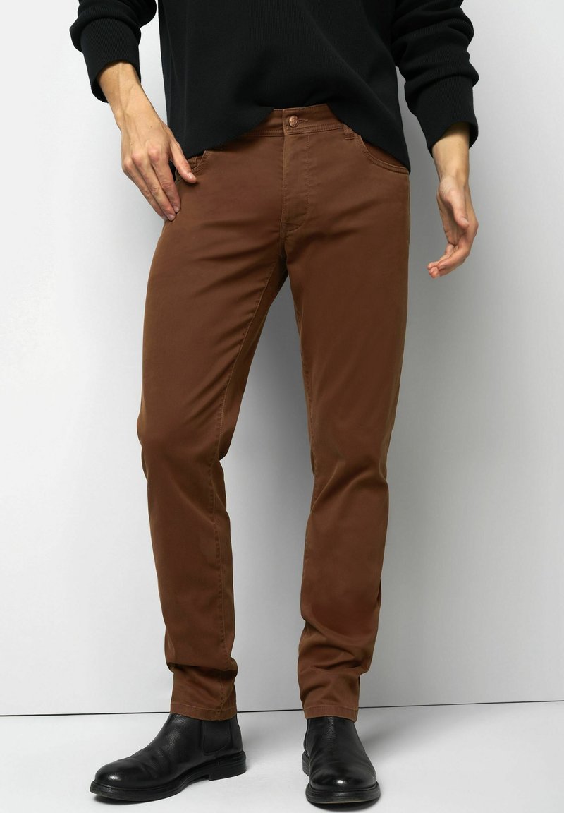 Man in bruin slim-fit broek en zwarte leren enkelboots, staand tegen een effen lichte achtergrond.