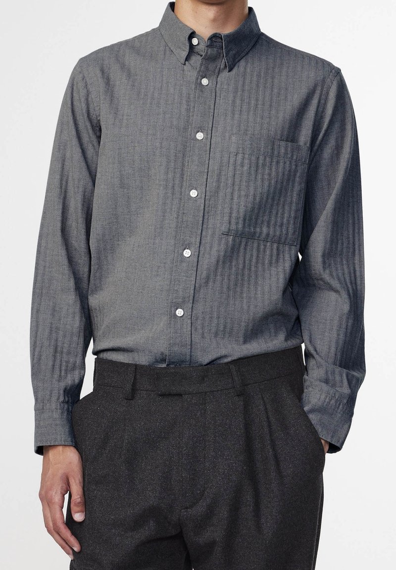 Grey button-down shirt featuring a vertical stripe pattern, a breast pocket, and a classic collar, paired with dark, tailored trousers.