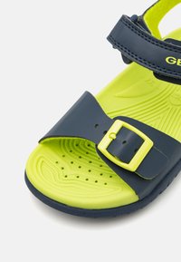 Sandalia azul marino con hebilla amarilla y plantilla verde lima. Presenta una superficie texturizada con perforaciones para mayor transpirabilidad.