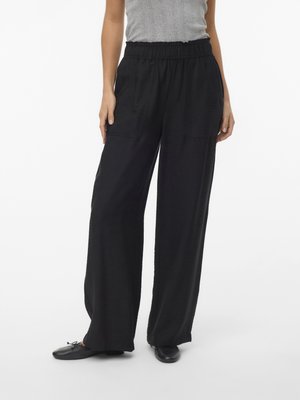 Donna che indossa pantaloni neri a gamba larga con fascia elastica in vita, una canotta grigia e ballerine nere, in piedi su sfondo bianco.