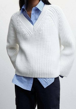 Person iført en hvid tekstureret V-hals trøje over en blåstripet skjorte med knapper og mørke bukser, stående med hænderne bag ryggen.