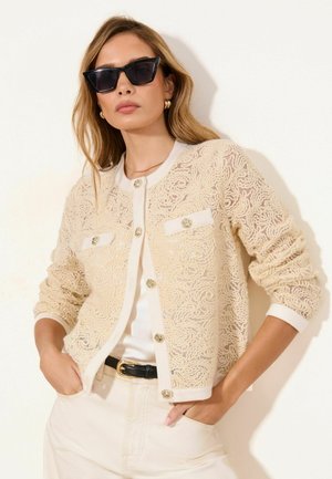 Kobieta w kremowym, teksturowanym kardiganie z złotymi guzikami, białym topie, kremowych spodniach, czarnym pasku, złotych kolczykach kołach i czarnych okularach przeciwsłonecznych w kształcie kocich oczu.