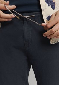 Marineblauwe broek met een gladde textuur, voorzien van een trekkoord met metalen accenten. Aan weerszijden zijn zijzakken zichtbaar.