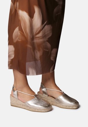 Espadrilles argentées métalliques avec une semelle en jute tressé, dos ouvert et lanières croisées autour de la cheville. Texture lisse avec un design à bout arrondi.