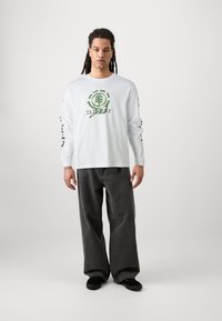 Camicia bianca a maniche lunghe con grafica frontale che presenta un design di un albero verde e testo, abbinata a pantaloni grigi a gamba larga e scarpe nere.