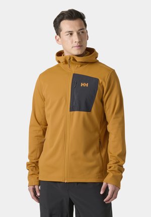 Hombre con sudadera con cremallera color mostaza y bolsillo negro en el pecho con logo, mirando hacia un lado sobre un fondo liso.