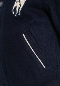 Marinblå ulljacka med dekorativ djurbroderi, med en sidficka kantad i en kontrasterande ljusare färg.