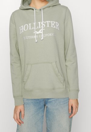 Personne portant un sweat à capuche Hollister vert clair avec des cordons blancs et une poche avant, associé à un jean bleu clair.