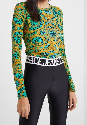 Vrouw draagt een turquoise top met lange mouwen met een patroon en gouden barokke ontwerpen, gecombineerd met zwarte legging met een witte tailleband van Versace Jeans Couture.
