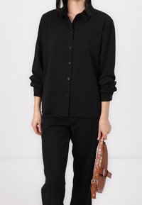 Camicia nera con bottoni, maniche arrotolate, texture liscia e colletto classico. Abbinata a pantaloni neri e una borsa in pelle stampata coccodrillo marrone.