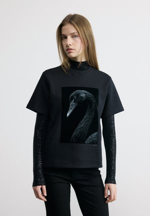 Νεαρή γυναίκα που φοράει μαύρα πουκάμισα με στρώσεις, ένα μακρυμάνικο και ένα κοντομάνικο, με το κοντομάνικο να έχει μεγάλο γραφικό μαύρου κύκνου στο μπροστινό μέρος.