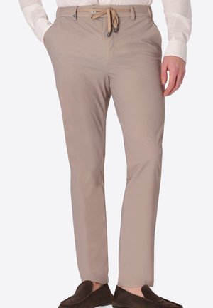 Uomo che indossa pantaloni beige slim-fit con coulisse e camicia bianca, abbinati a scarpe slip-on marrone scuro, in piedi con le mani vicino alle tasche.