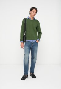 Zelený sveter s pleteným vzorom cez modrú kockovanú košeľu, v kombinácii so svetlomodrými džínsami a čiernymi mokasínami. Nosená je čierna crossbody taška.