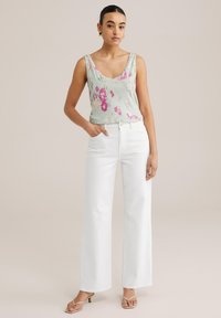 Cvetasti, svetlo zeleni top s tankimi naramnicami in v-izrezom; kombiniran s širokimi belimi kavbojkami; model stoji z roko v žepu.