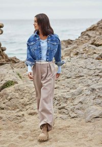 Chaqueta de denim, top de canalé azul claro, pantalones beiges con pliegues y botas de tobillo marrones. Entorno natural con rocas y una playa de arena al fondo.