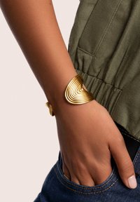 Bracciale a manica in tono oro con un design ondulato e rigato. Finitura metallica liscia; mette in evidenza una forma unica. Indossato su un polso con tono di pelle chiaro.