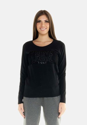 Jonge vrouw met lang bruin haar glimlacht, draagt een zwart shirt met lange mouwen met de tekst "LEONE 1947" en grijze broek, staand tegen een witte achtergrond.