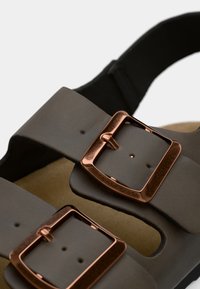 Brown sandals with two wide straps and square copper buckles, featuring a textured finish and a cushioned footbed with a neutral color.