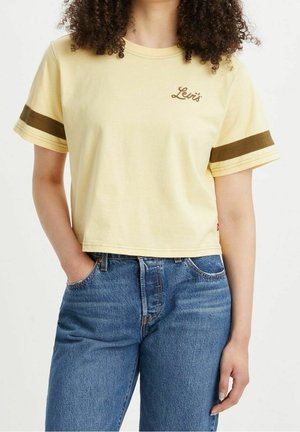 Vrouw draagt een cropped geel Levi's T-shirt met bruine strepen op de mouwen en blauwe jeans, staand met één hand in de zak.