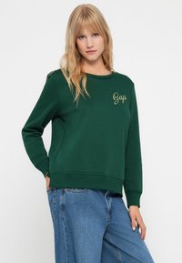 Sudadera verde bosque con cuello redondo y puños acanalados. Presenta el logo dorado de "Gap" en el lado izquierdo del pecho. Fabricada con una tela suave y texturizada.