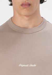 Beige kortermet t-skjorte med rund hals laget av myk stoff med strukturert ribbestrikket krage; har hvit brodert tekst nederst.