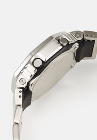 Montre en acier inoxydable argenté avec une finition brossée, accents en caoutchouc noir et un boîtier rond. Dispose de plusieurs boutons et d'un bord lisse.