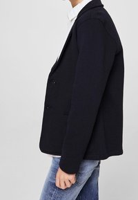 Tmavě modrý blejzr z texturované látky s přizpůsobeným střihem, vybavený klopovým límcem s výstřihem do V a dvěma knoflíky. Nošený přes bílou košili a džíny.