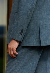 Traje de lino azul grisáceo con textura visible, que presenta cuatro botones negros en el puño y una pequeña abertura en la parte trasera de la chaqueta.