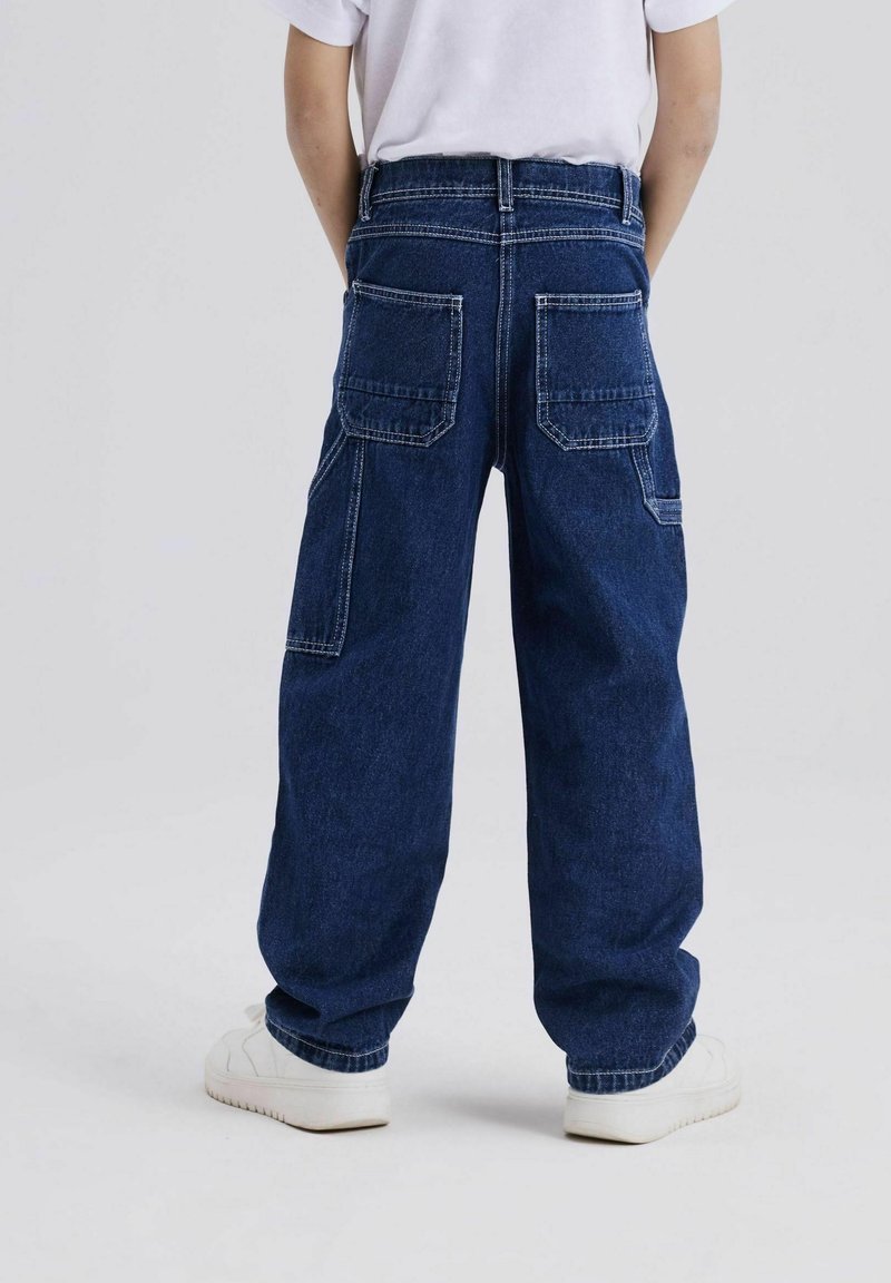 Blauwe denim broek met een relaxte pasvorm, voorzien van twee achterzakken en witte stiksels. Gecombineerd met witte sneakers, gezien vanuit de achterzijde.